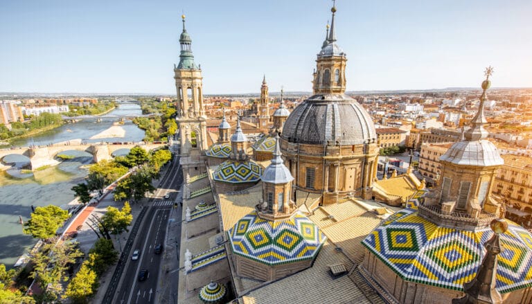 View of Zaragoza's iconic Basilica del Pilar with colorful tiled domes and the Ebro River in the background, showcasing the architectural beauty of Spain.