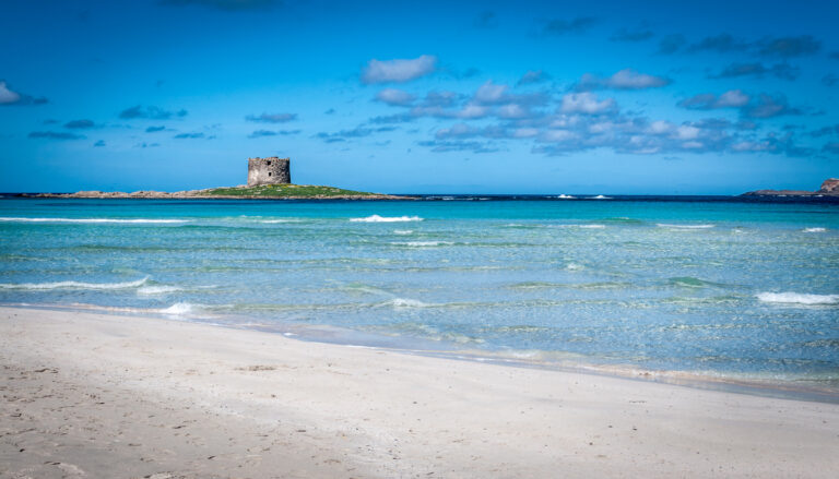 A stunning beach in Sardinia, Italy, with crystal-clear turquoise waters, a sandy shore, and a historic stone tower on a distant island. The blue sky with scattered clouds completes the serene and picturesque scene.