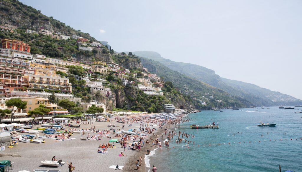A bustling beach in Positano, Italy, with numerous people lounging, swimming, and enjoying the sunny weather, while colorful buildings cascade down the hillside behind them. A prime example of the Best Beaches in Italy.