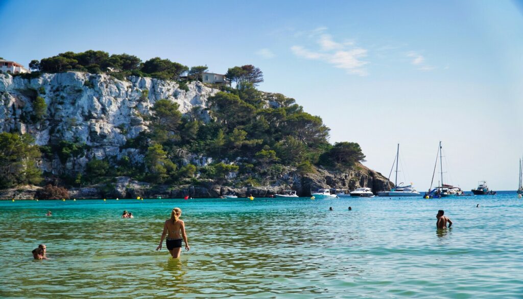 Visitors enjoying the crystal-clear waters of Cala Galdana, one of the best beaches in Spain, with boats anchored nearby and lush greenery covering the white cliffs.