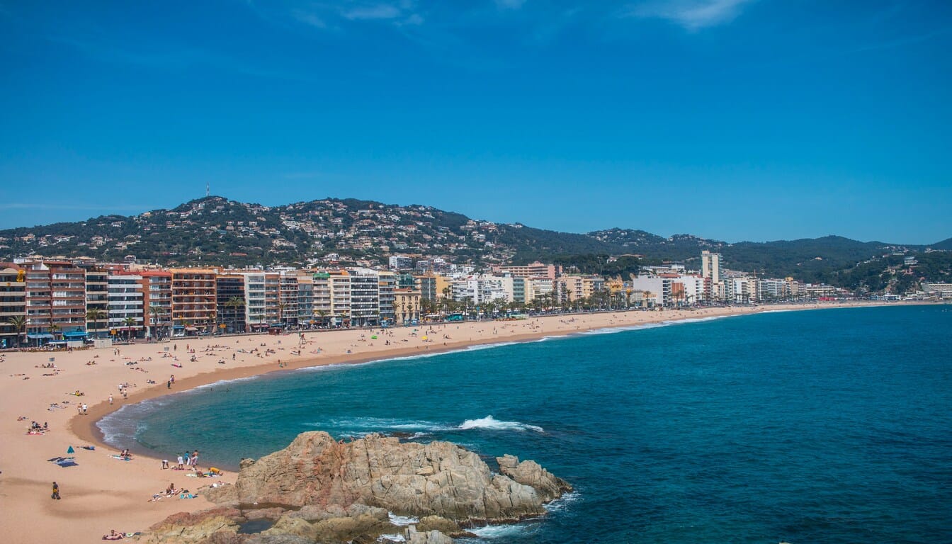 Panoramic view of Lloret de Mar Beach, one of the best beaches in Spain, featuring golden sands, vibrant blue waters, buildings along the shoreline, and green hills in the background.